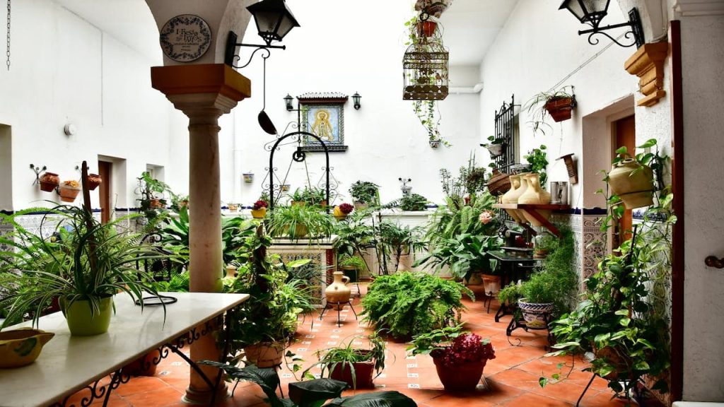 Traditional Spanish-style courtyard with white walls, terracotta floors, ornate column, wrought iron lanterns, and abundant potted plants throughout