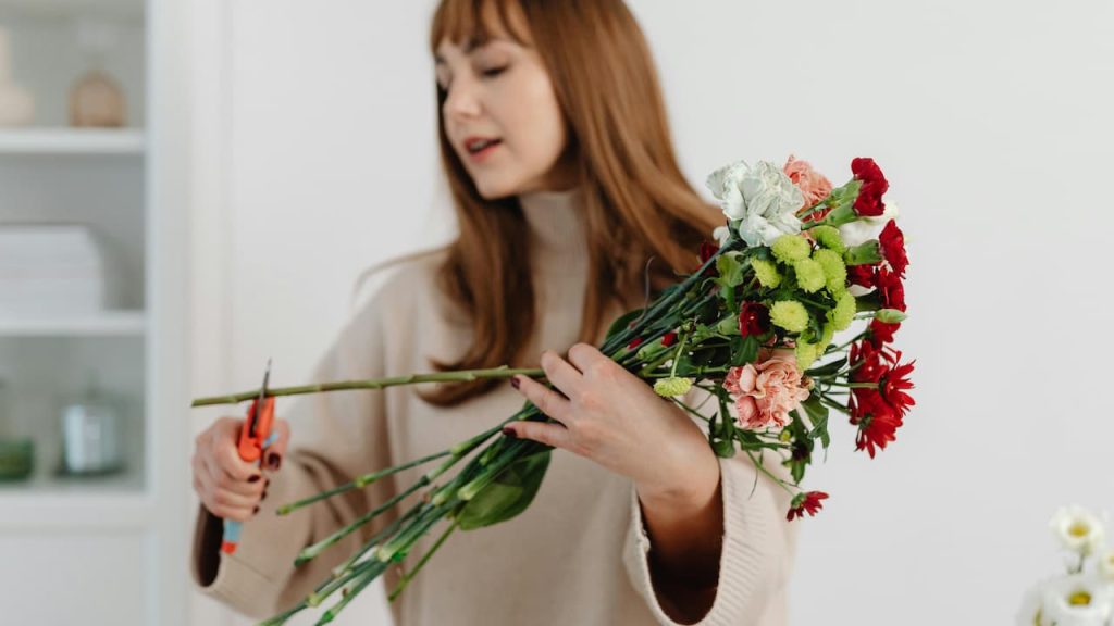 Woman with blonde hair cutting flowers with a pair of scissors, holding freshly harvested blooms, outdoor garden setting, focused on hands and flowers