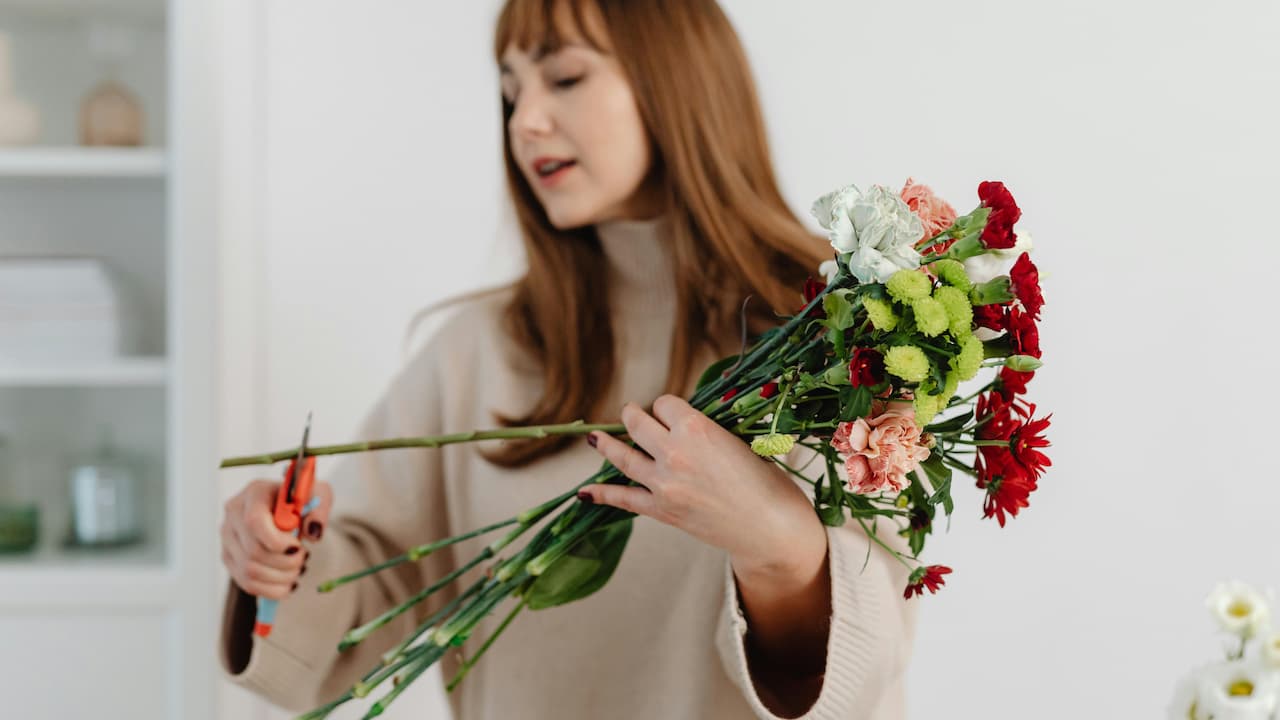 Woman with blonde hair cutting flowers with a pair of scissors, holding freshly harvested blooms, outdoor garden setting, focused on hands and flowers