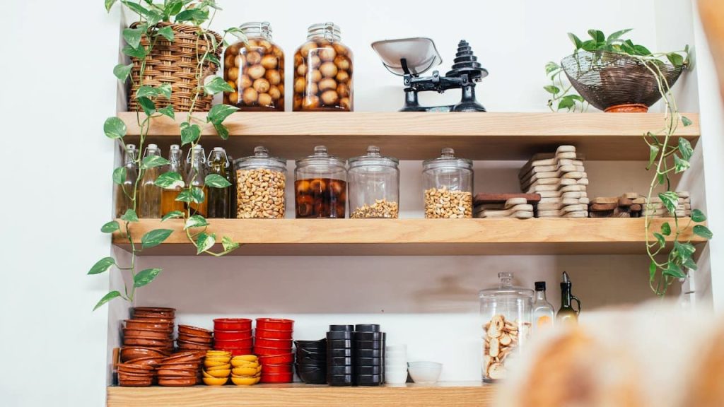 Organized kitchen shelves displaying glass jars with stored food, colorful bowls, vintage scale, and trailing green plants