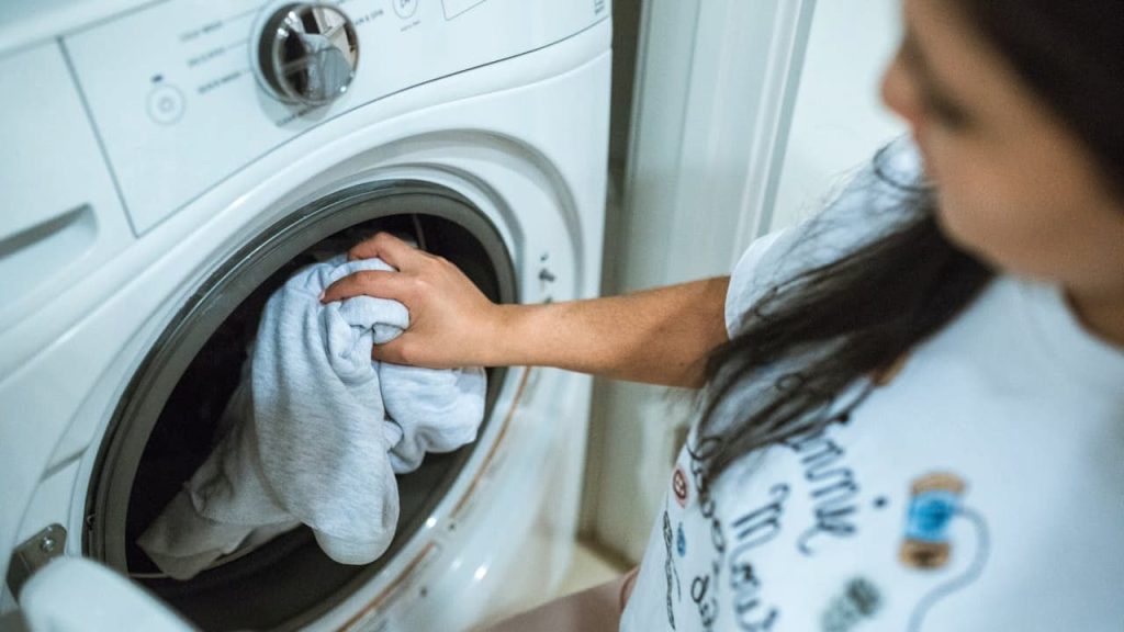 Woman loading white towels into front-loading washing machine in bright laundry room with white cabinets and tiled floor