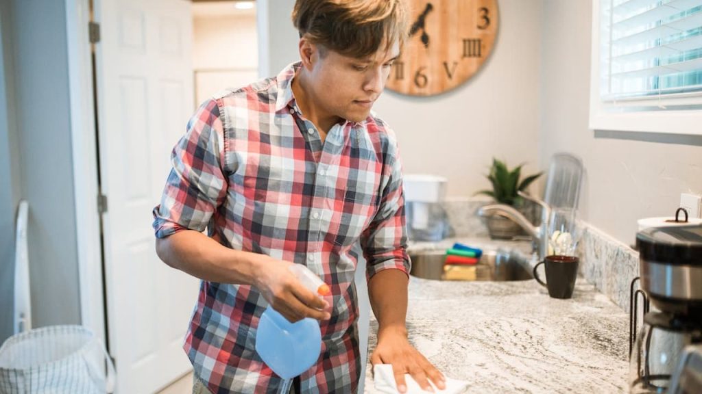 Man in plaid shirt cleaning kitchen counter with blue spray bottle in modern kitchen with granite countertops