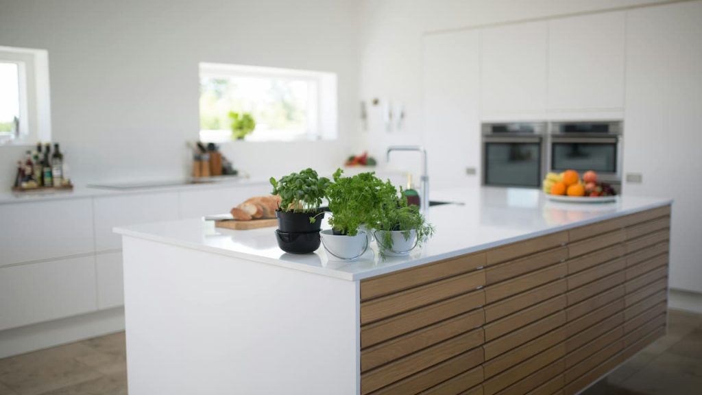 Modern white kitchen with wooden island drawers, featuring potted herbs, built-in ovens, and minimalist design with natural lighting