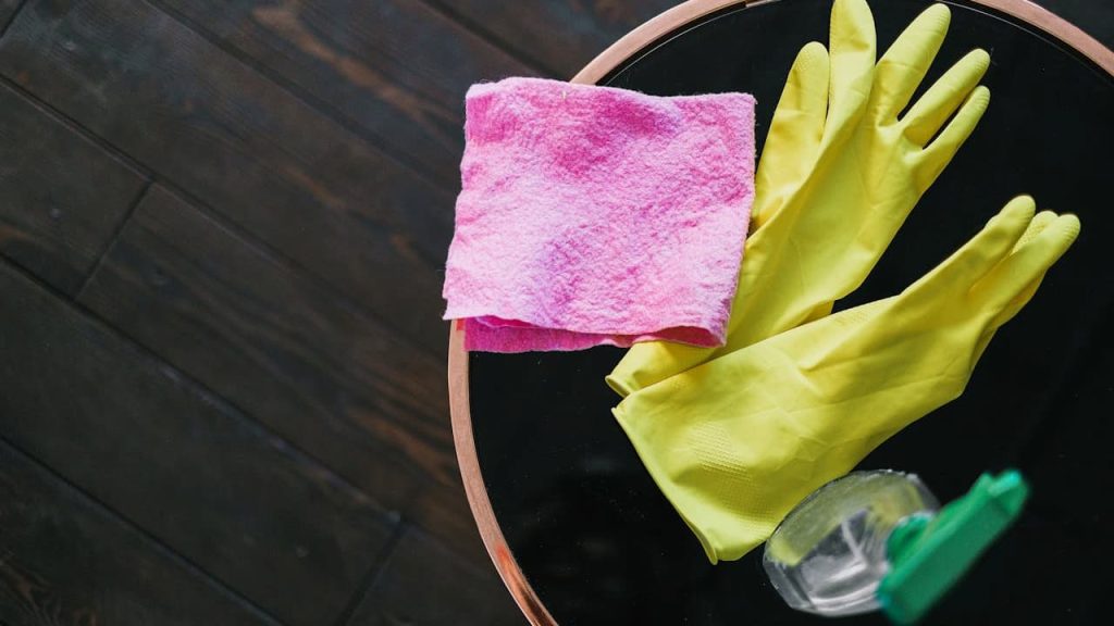 Black bowl containing yellow rubber gloves, pink cleaning cloth, and spray bottle on dark wooden floor surface