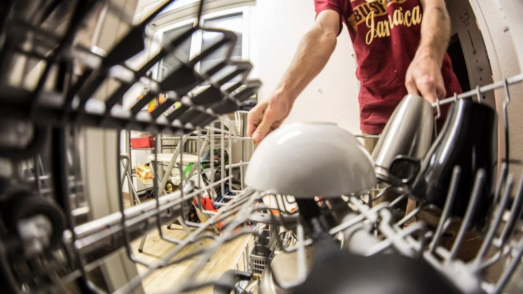 Person in red shirt loading white plate into open dishwasher with metal racks and various dishes inside kitchen