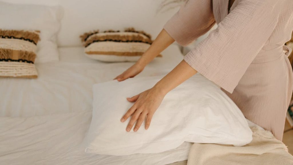 Person in beige sweater making bed with white linens, smoothing bedding with hands in neutral bedroom