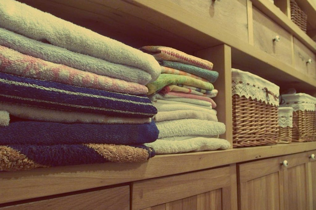 Neatly folded towels and linens stacked on wooden shelving unit, wicker baskets with lace trim on right side, organized linen closet storage