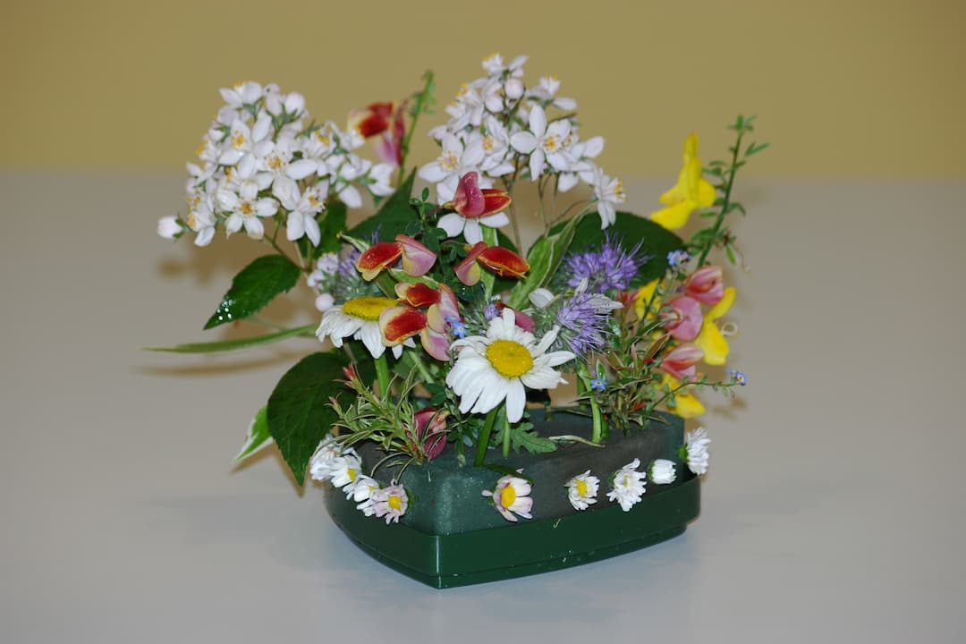 Small floral arrangement featuring daisies, chive blossoms, and various wildflowers inserted into a green floral foam base