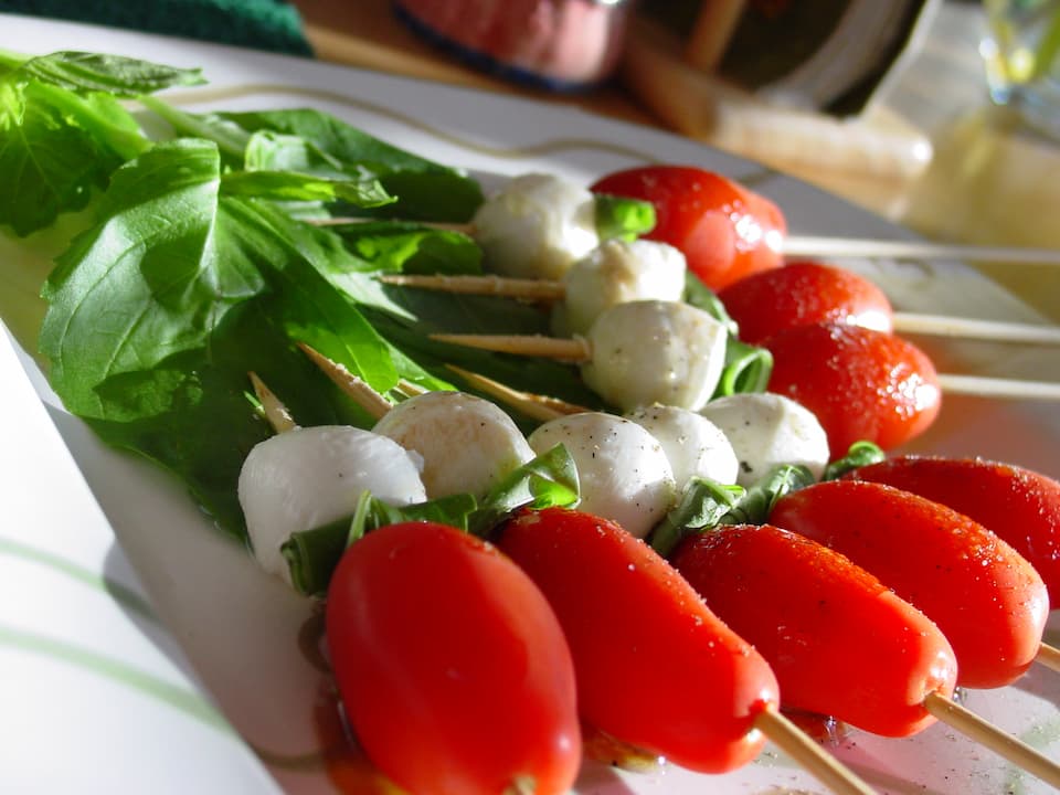 Caprese salad skewers with cherry tomatoes, mozzarella balls, and fresh basil leaves on toothpicks, served on a white plate with leafy greens