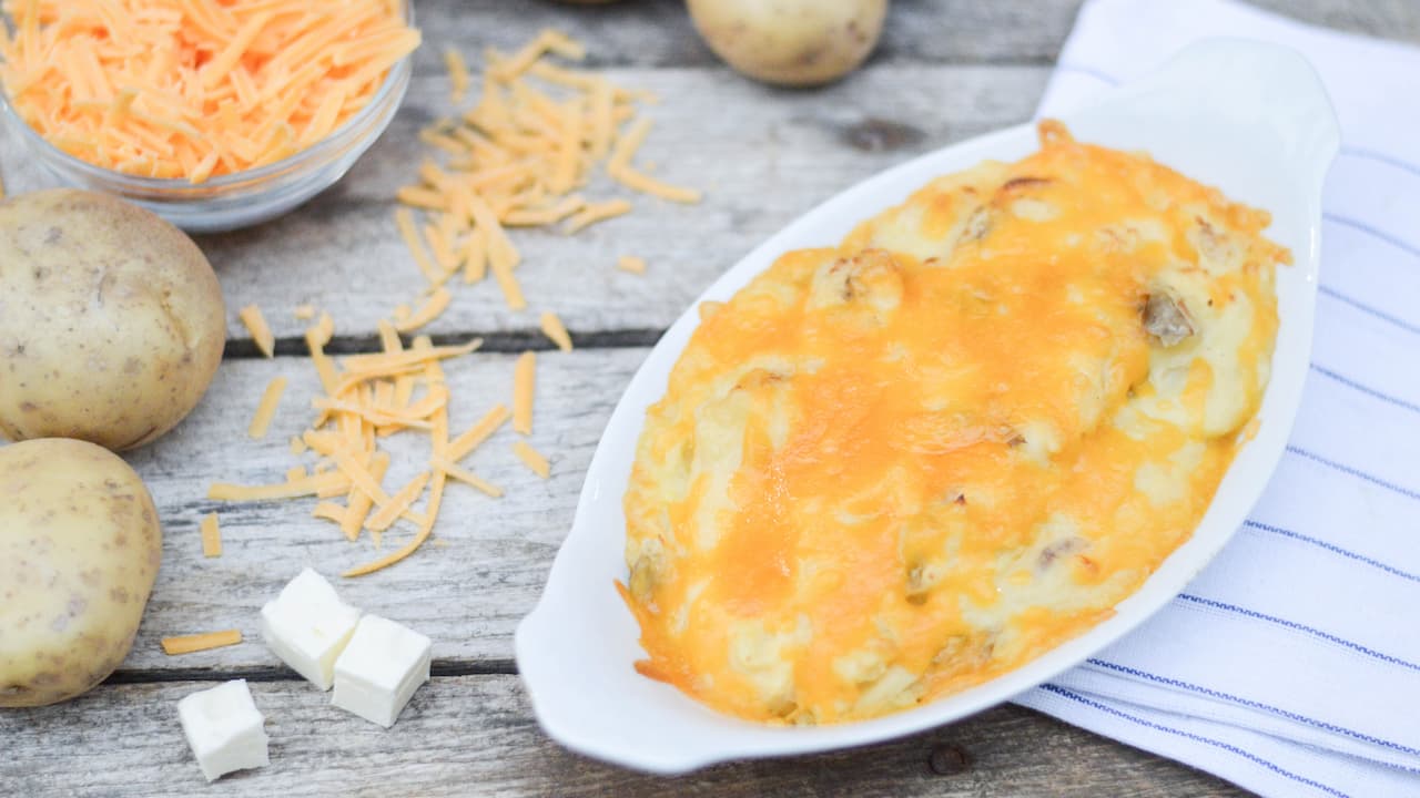 White oval dish filled with cheesy mashed potatoes topped with melted cheddar, surrounded by raw potatoes, shredded cheese, and butter cubes on a rustic wooden surface