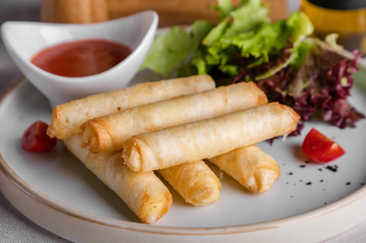 Crispy spring rolls served on a white plate with dipping sauce, fresh lettuce, and cherry tomatoes