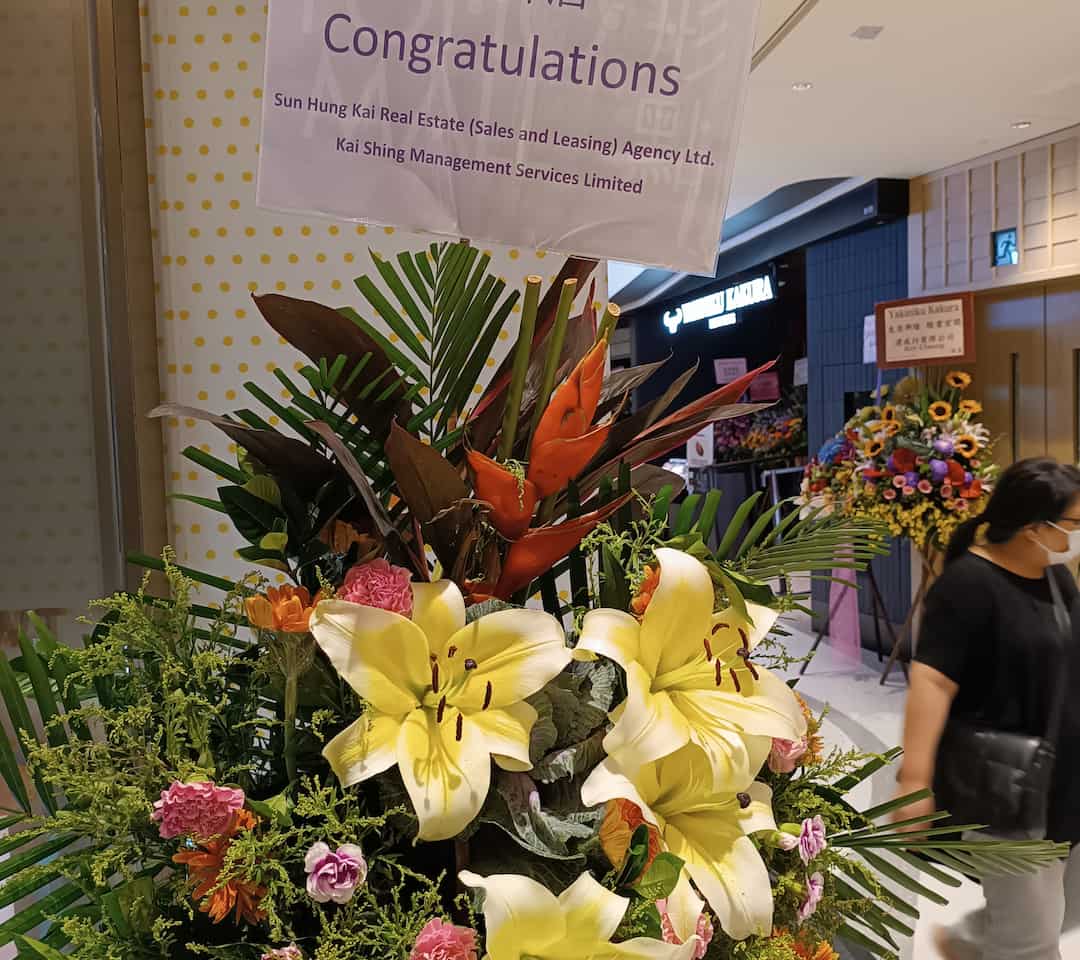 Mixed floral arrangement with large yellow lilies, orange bird-of-paradise flowers, pink and orange carnations, assorted greenery, and palm leaves, placed below a congratulatory sign in a shopping mall