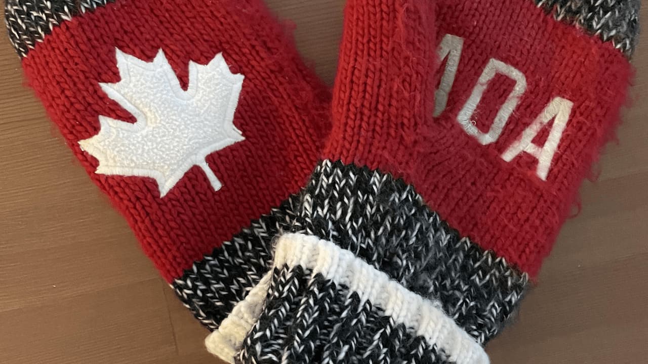 Pair of red, black, and white knit wool mittens with a white maple leaf on one mitten and the word "CANADA" partially visible on the other, placed on a wooden surface