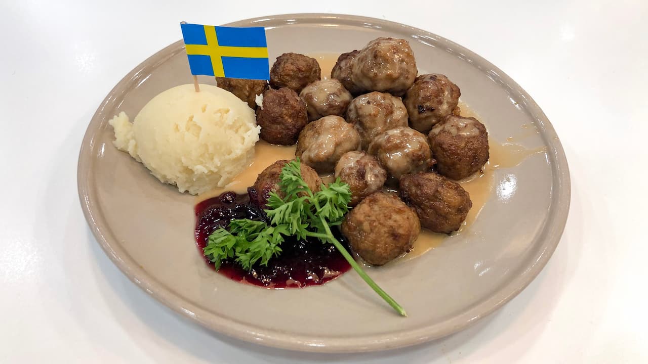 Plate of Swedish meatballs served with brown gravy, a scoop of mashed potatoes, lingonberry sauce, and a garnish of parsley, topped with a small Swedish flag