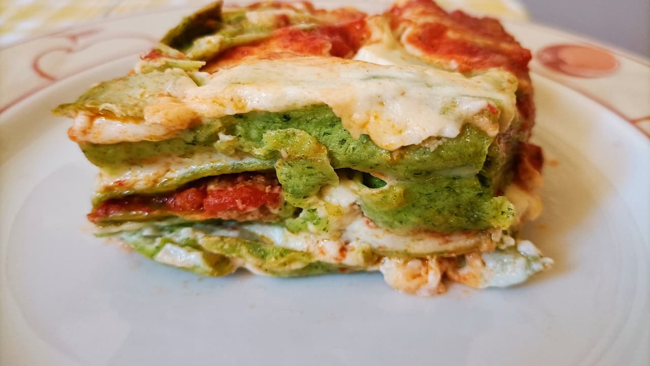 Slice of green pesto lasagna with layers of spinach pasta, tomato sauce, melted cheese, and béchamel, served on a white plate with a decorative rim