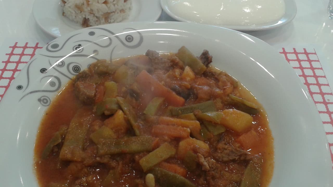 Plate of orzo stew with green beans, carrots, potatoes, and beef in a tomato-based broth, served with a side of rice and a bowl of yogurt on a red-checkered placemat