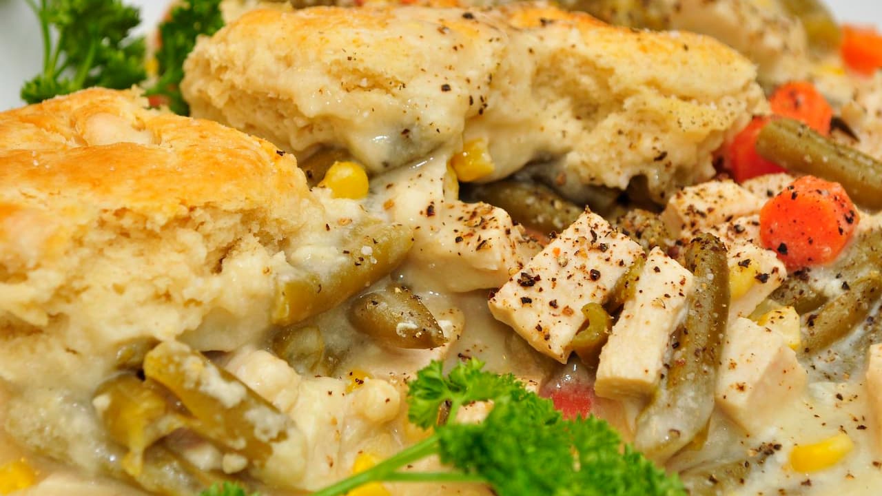 Close-up of a chicken pot pie with golden biscuit topping, diced chicken, green beans, carrots, corn, and creamy sauce, garnished with fresh parsley