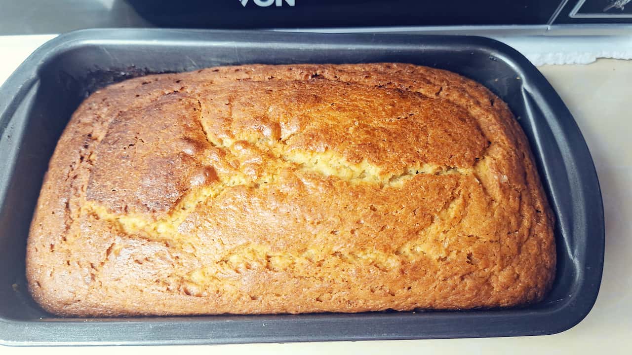 Freshly baked golden-brown banana bread in a rectangular black non-stick loaf pan, resting on a kitchen countertop with an oven in the background