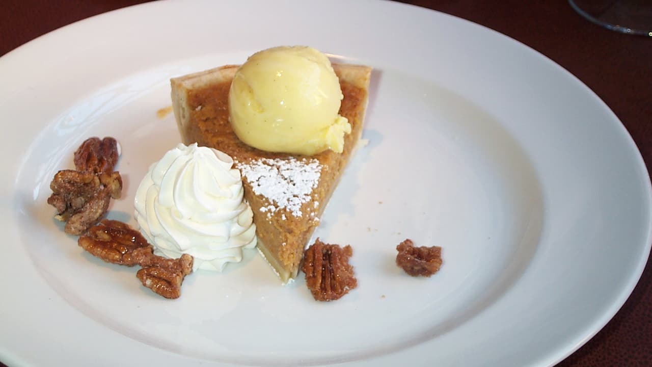 Slice of sweet potato pie topped with a scoop of vanilla ice cream, garnished with whipped cream, powdered sugar, and candied pecans, served on a white plate