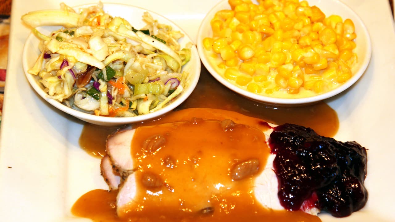 Plate with roasted turkey slices topped with brown gravy and cranberry sauce, served with bowls of creamed corn and colorful coleslaw