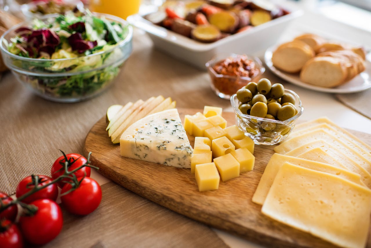 wooden cheese board with sliced cheeses, cubed cheese, apple slices, green olives, surrounded by salad, bread, tomatoes, and roasted vegetables
