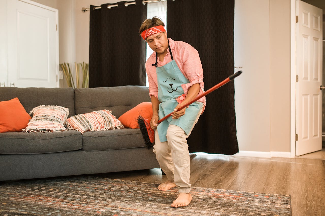 man barefoot in living room, wearing red bandana and blue apron, sweeping rug with red broom near gray couch