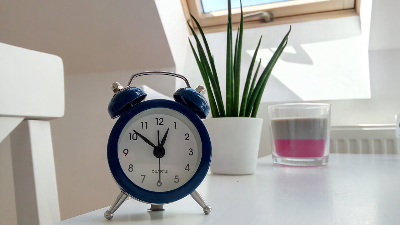 blue analog alarm clock on white table, green potted plant, layered candle in glass, daylight through skylight