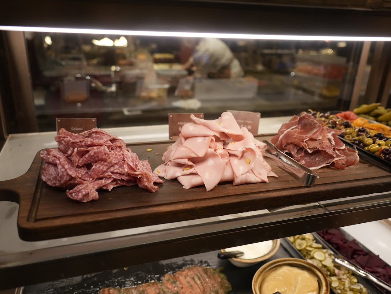 wooden board with piles of salami, mortadella, and prosciutto, set on buffet counter with olives and pickles nearby