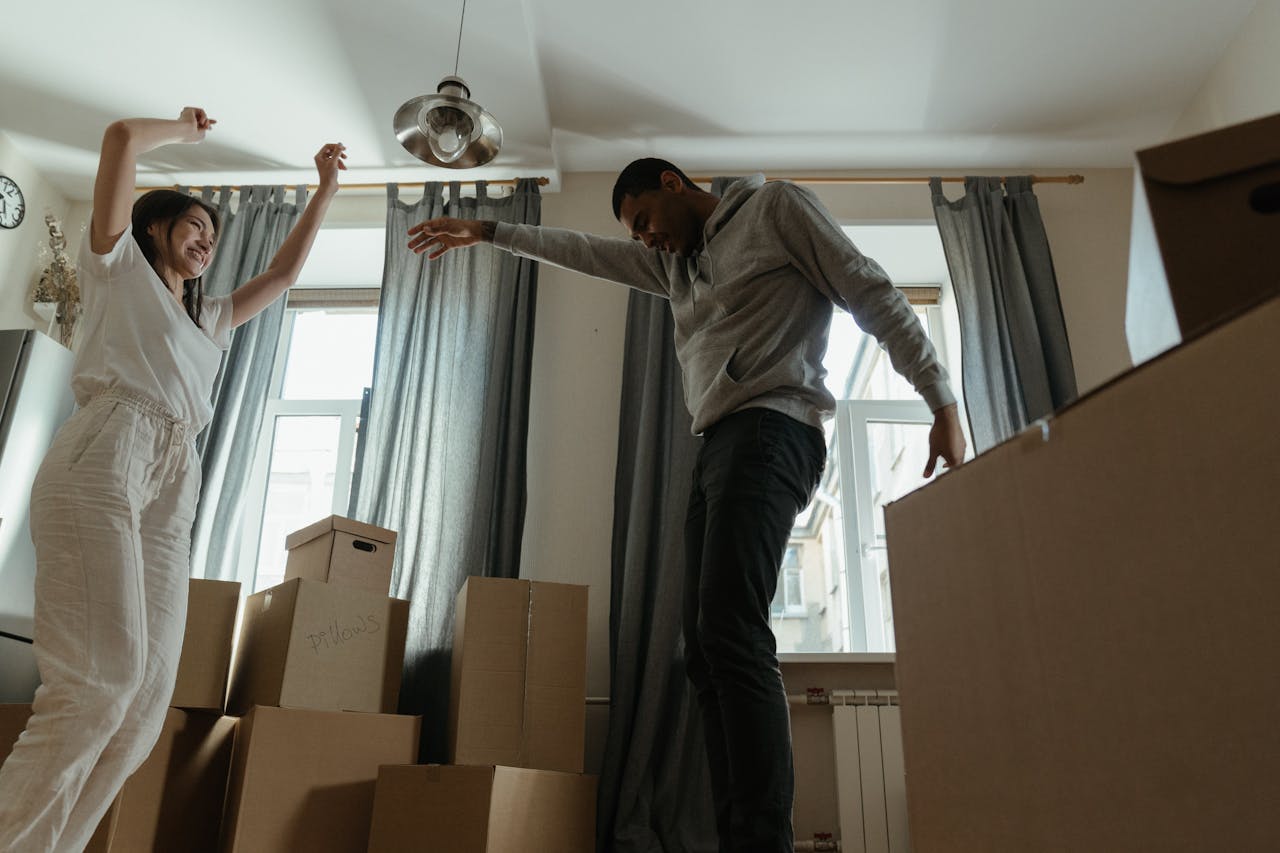 smiling couple dancing between moving boxes in bright room with curtains, celebrating new home move-in day