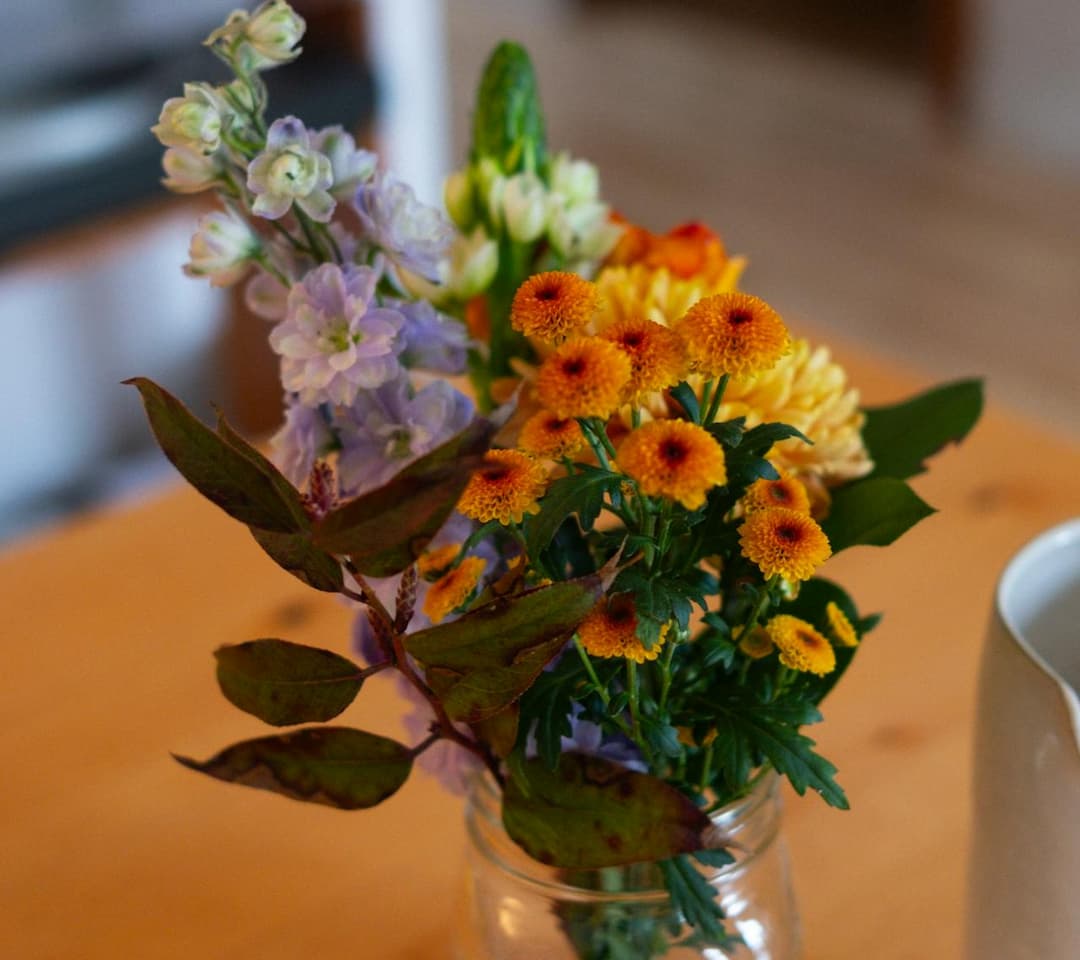 Colorful bouquet of fresh flowers, including orange chrysanthemums and lavender blooms, arranged in a mason jar on a wooden table
