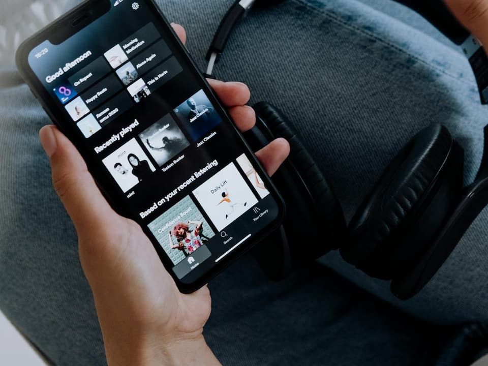 Person holding a smartphone with a music streaming app open, showing playlists and recently played songs, next to a pair of black headphones resting on denim-clad legs