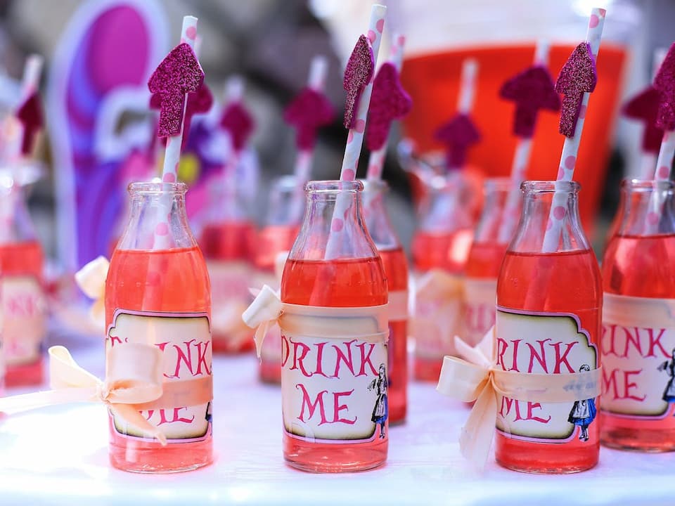 Glass bottles filled with pink drink, each labeled “Drink Me” and decorated with ribbon and glittery pink umbrella straws, arranged on a white table