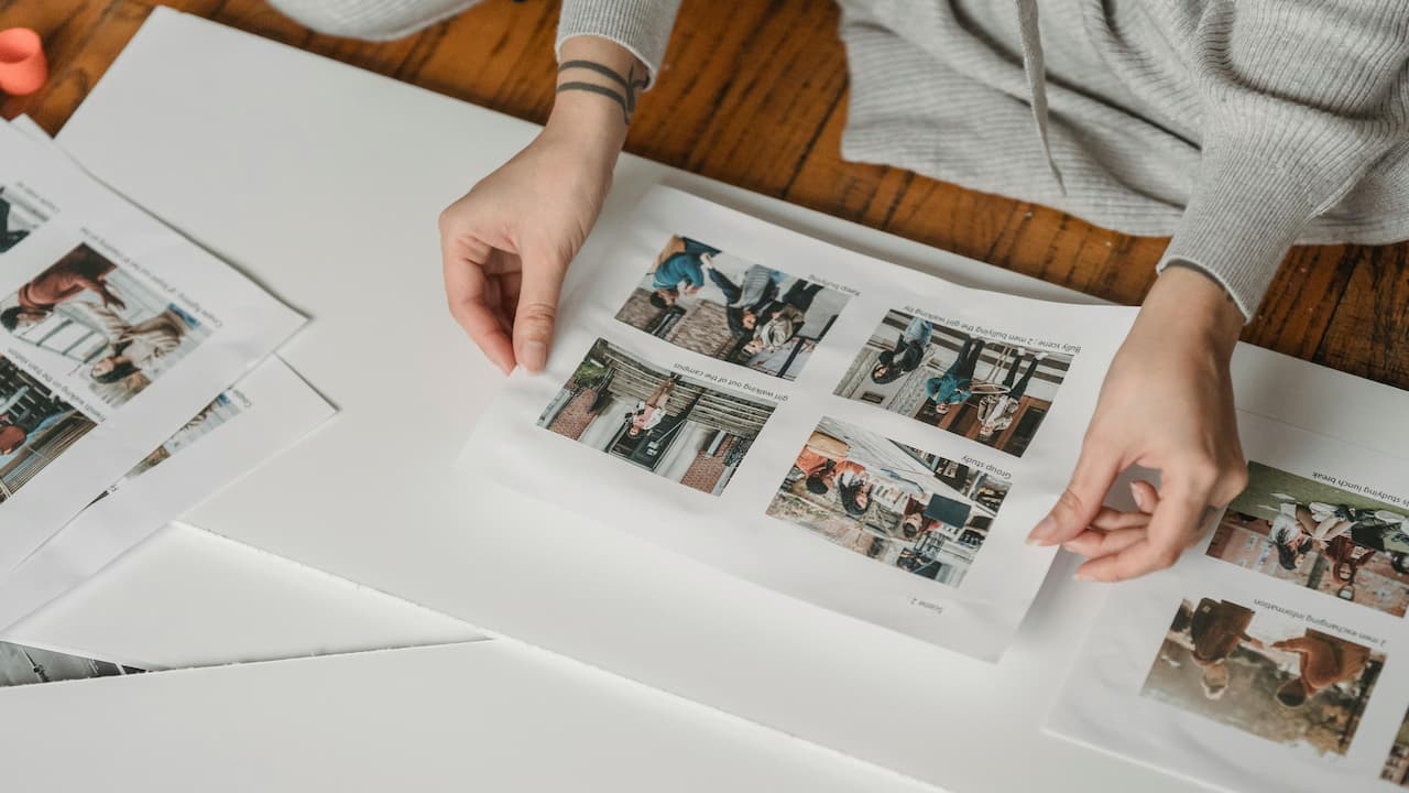 Person in grey clothing assembling a DIY photo album, arranging printed photo sheets with multiple images onto white cardstock on a wooden floor