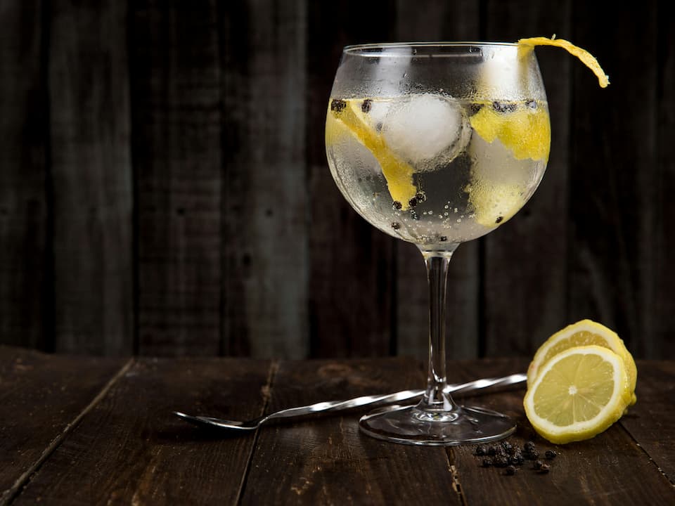 Gin and tonic served in a stemmed glass with ice cubes, lemon peels, and peppercorns, placed on a rustic wooden surface beside a lemon slice and a twisted cocktail spoon