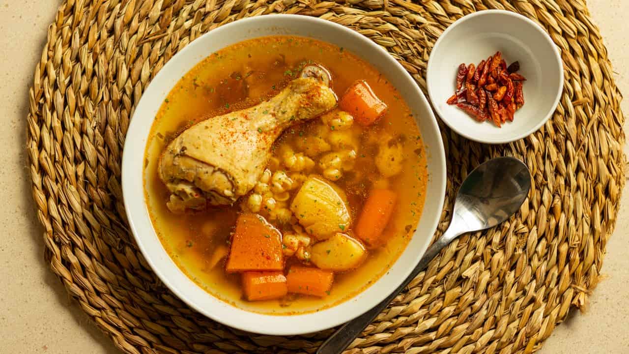 Bowl of chicken stew with a drumstick, diced carrots, potatoes, and hominy in a rich broth, served on a woven placemat with a side dish of dried chili peppers and a spoon