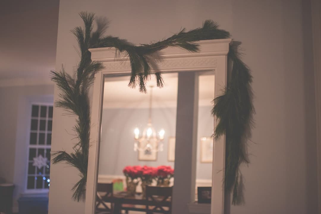 Mirror decorated with a pine garland, white frame, soft indoor lighting, reflection of chandelier, dining table with red poinsettia flowers, light blue walls, cozy holiday setting