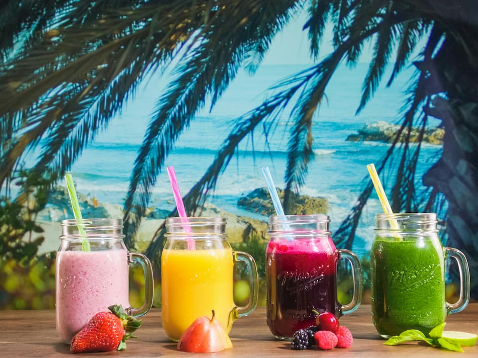 Four colorful smoothies in mason jar mugs with straws pink, yellow, purple, and green arranged on a wooden surface with fresh fruit in front