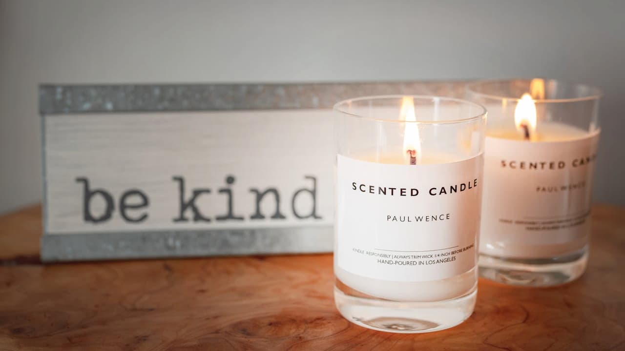 Two lit scented candles in clear glass containers labeled "Scented Candle Paul Wence," placed on a wooden surface with a decorative sign in the background that reads "be kind."
