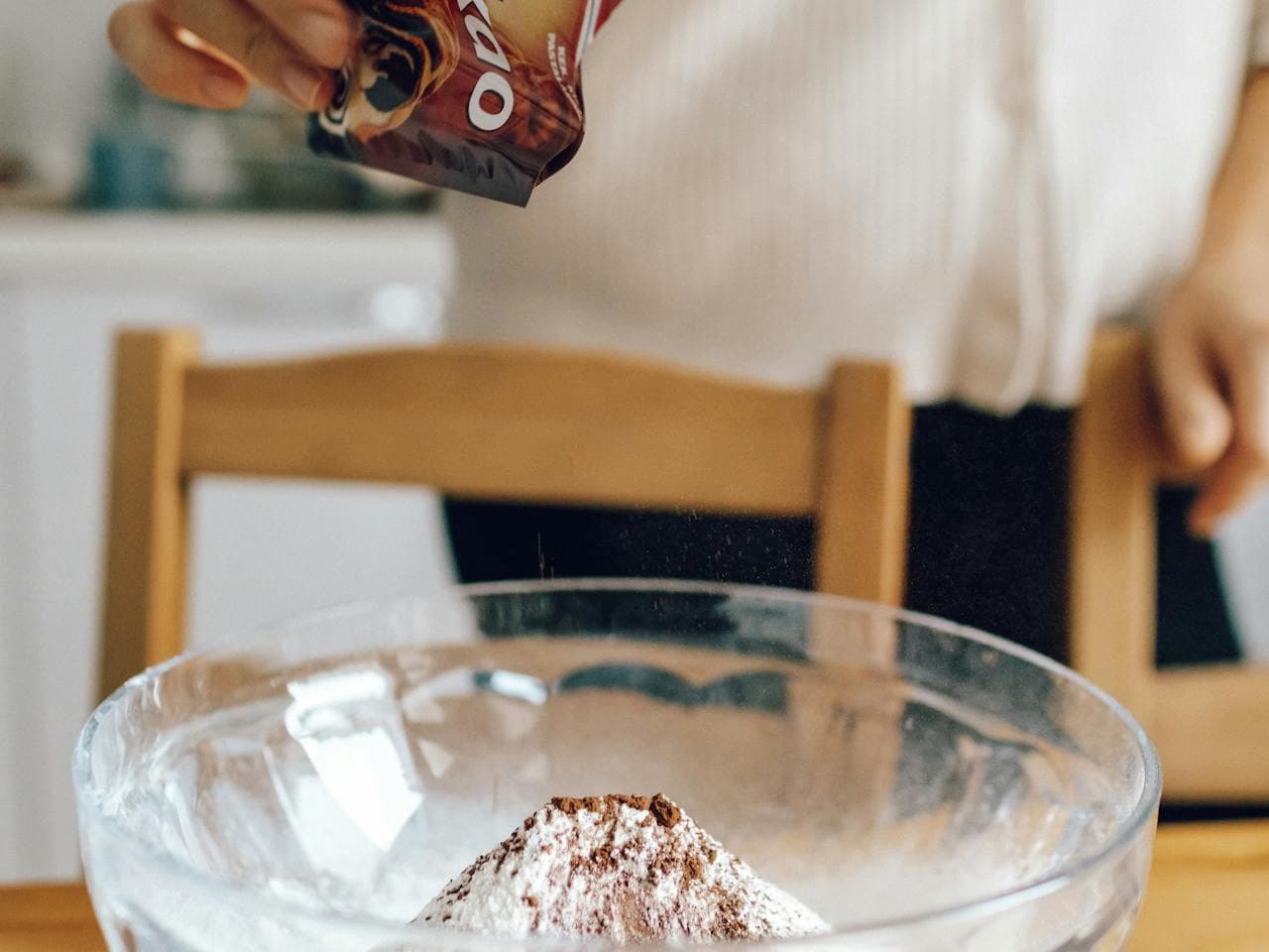 Person pouring cocoa powder from an Ovaltine packet into a glass mixing bowl filled with flour