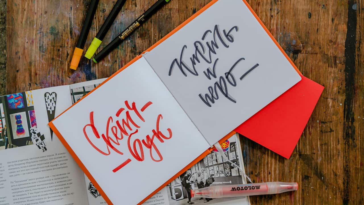 Open sketchbook with stylized Russian calligraphy in red and black ink, surrounded by highlighters, a red envelope, and art magazines on a rustic wooden table