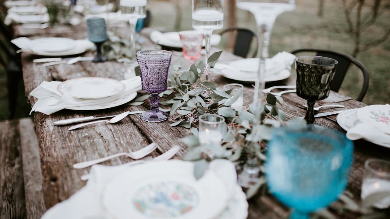 Rustic outdoor dining setup, long wooden table, white plates with floral patterns, folded napkins, assorted vintage-style glassware in blue, purple, and green, greenery centerpiece, metal chairs, soft natural lighting, relaxed and elegant atmosphere
