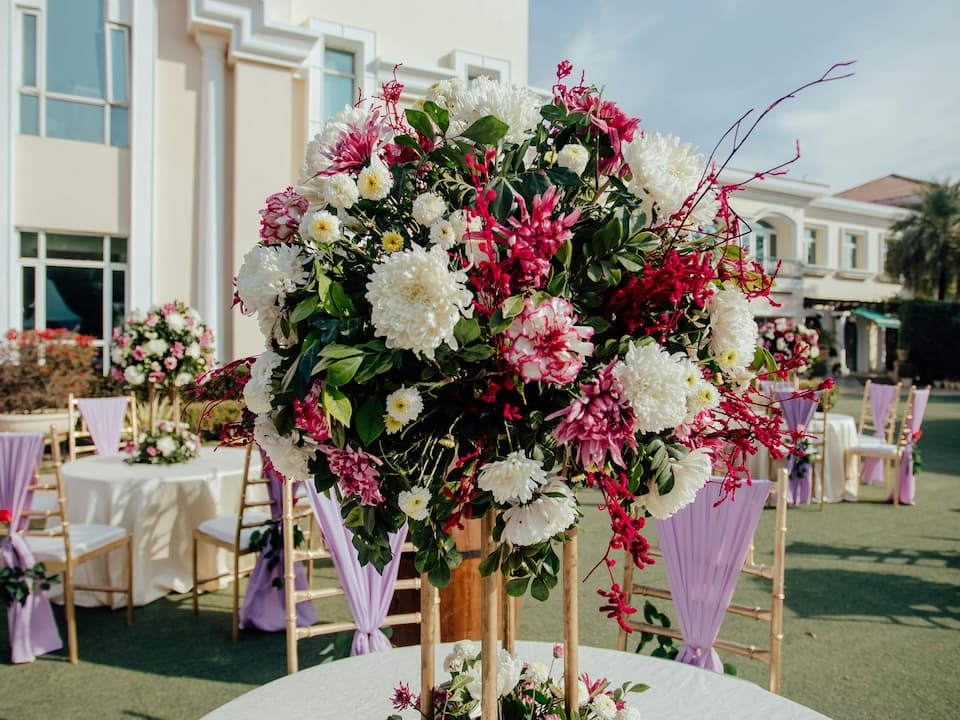 Outdoor wedding reception setup with a tall floral centerpiece of white and pink flowers and greenery on a round table, surrounded by gold chairs draped in lilac fabric