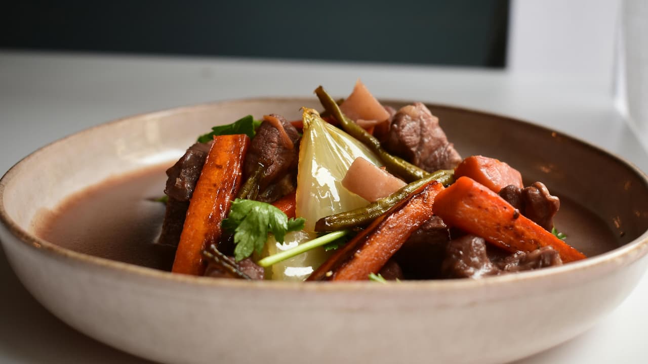 Beef stew in shallow bowl, chunks of beef, roasted carrots, green beans, onion wedge, herbs, light broth base, white and dark background contrast