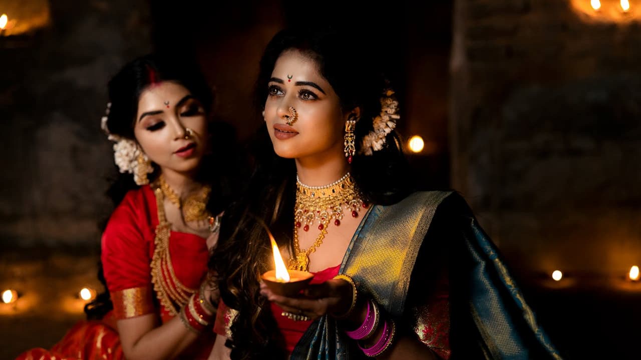 Two women dressed in traditional Indian attire, adorned with gold jewelry and silk-thread earrings, holding a lit oil lamp in a dimly lit festive setting