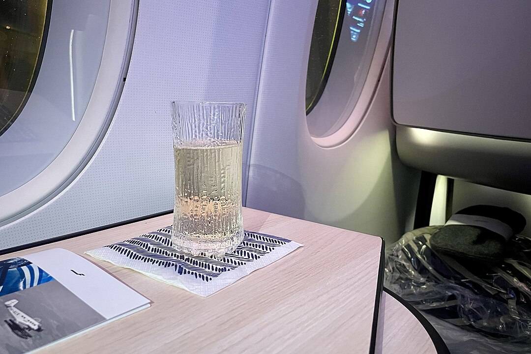 Glass of sparkling drink on a wooden airplane tray table, placed on a patterned napkin, seat and window visible in the background, soft cabin lighting, premium or business class airline setting