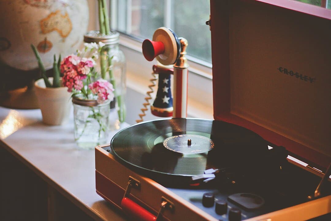 vinyl record playing on red Crosley turntable, vintage patriotic telephone nearby, pink flowers in mason jars, globe and potted plant on windowsill, soft natural light