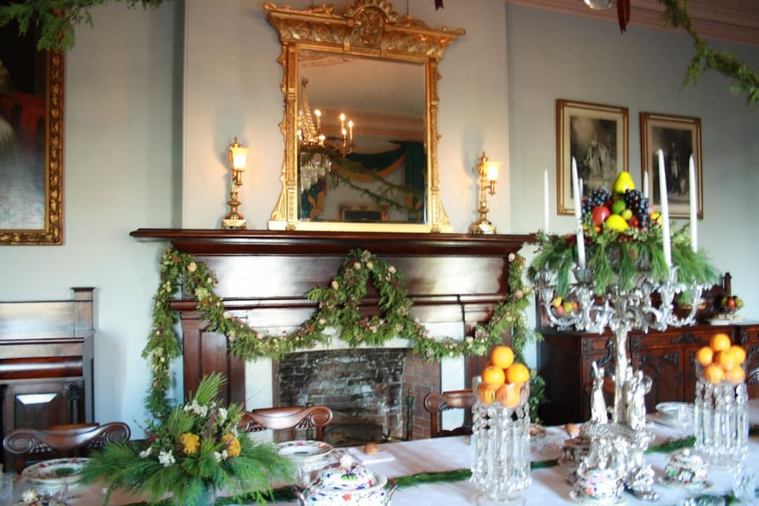 Festively decorated dining room with garland-draped fireplace mantle, ornate gold-framed mirror, crystal candleholders with fruit and greenery, formal dining table set with fine china and silverware, classical portraits on walls, chandelier reflection