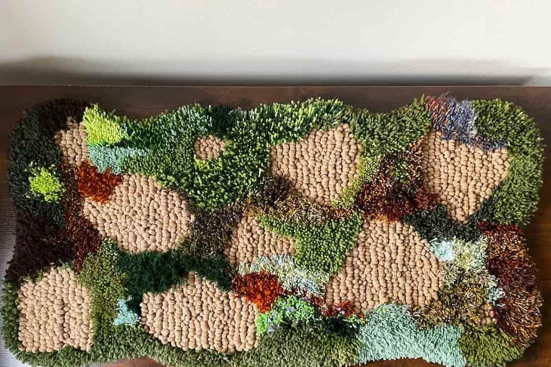 Textured handmade moss mat with varied tufted yarns in shades of green, brown, and earthy tones, mimicking a natural landscape with patches of moss, soil, and foliage
