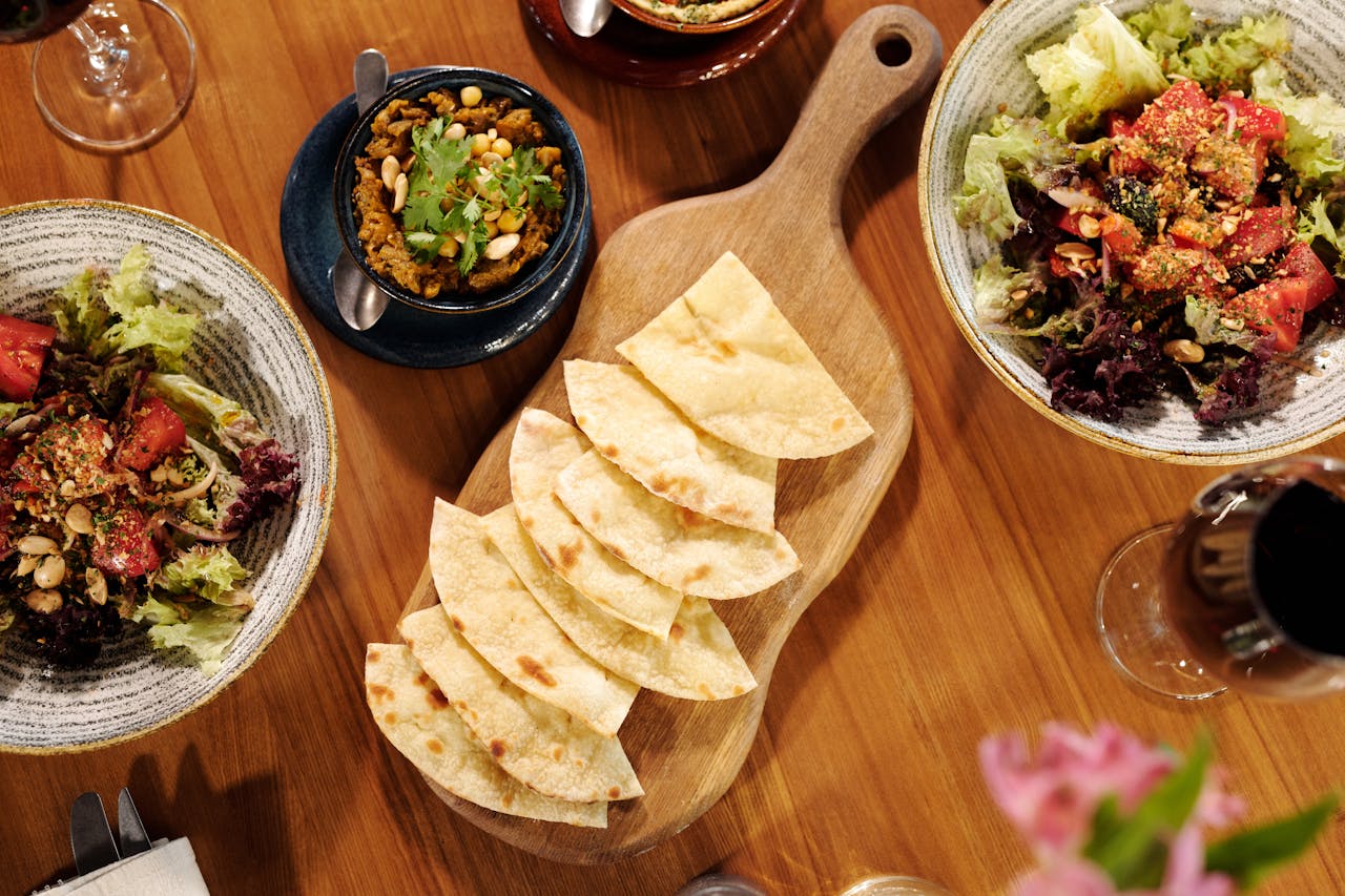 Pita chips arranged on a wooden board, surrounded by salads and dip bowls on a wooden table
