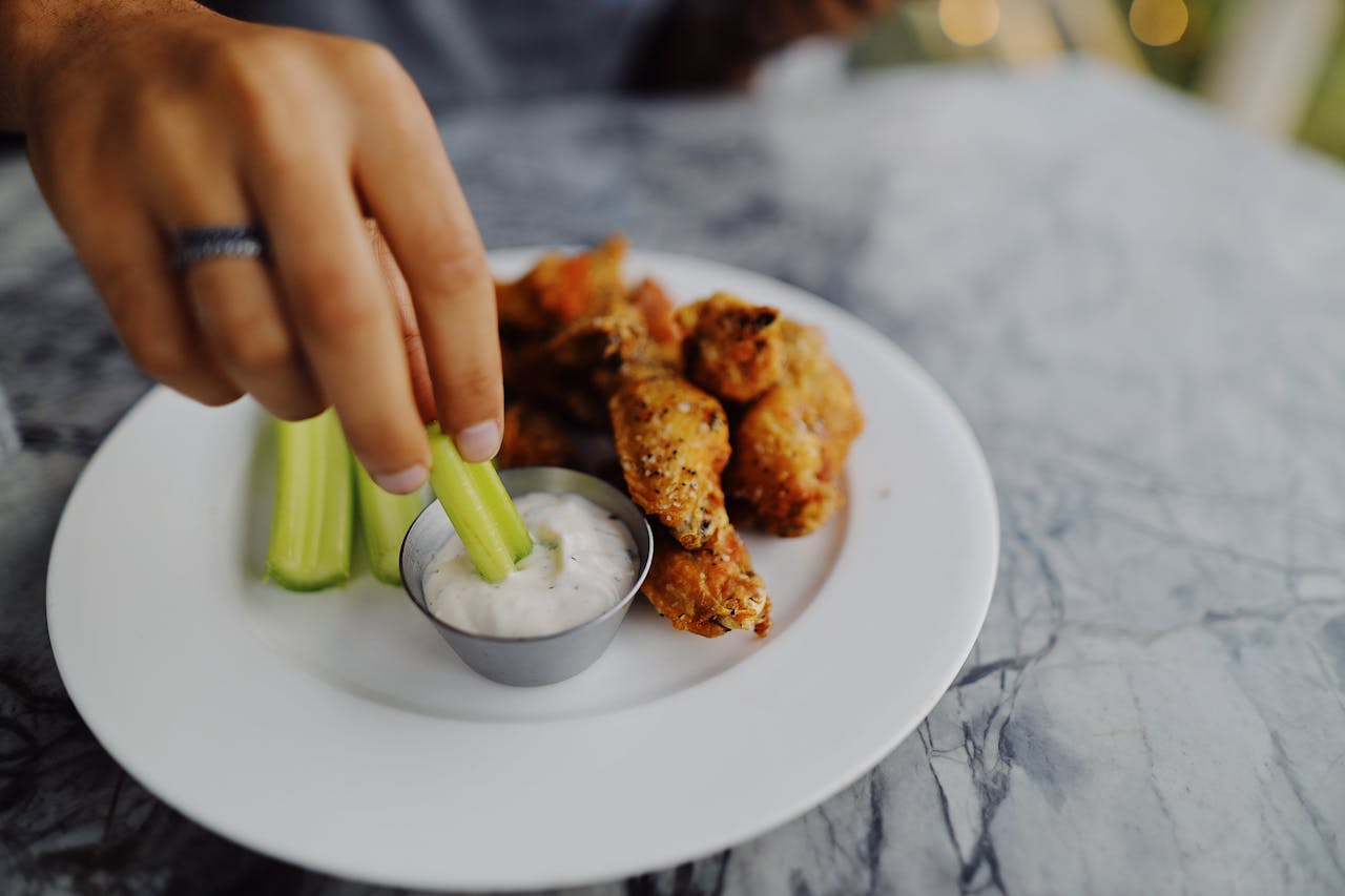 Hand dipping celery stick into ranch dressing, plate with chicken wings and celery on marble table